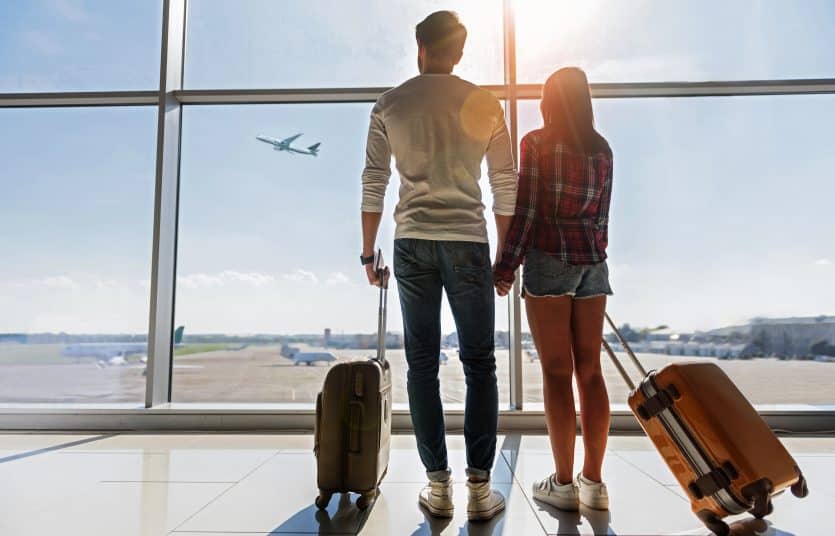 We are ready for new future. Young man and woman are watching flight at airport. They are standing and carrying luggage