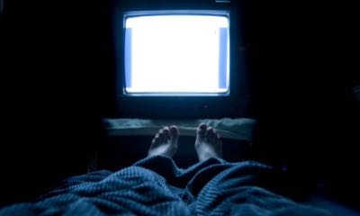 Person watches TV at night in his bed with his feet sticking up out of the blankets. The TV screen is blank white