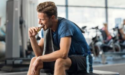 Sweaty young man eating energy bar at gym. Handsome mid guy enjoying chocolate after a heavy workout in fitness studio. Fit man biting a snack and resting on bench.