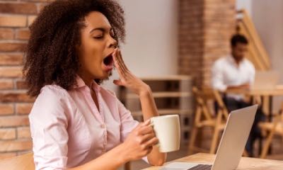 Young beautiful Afro-American businesswoman using laptop, holding cup and yawning while working in cafe