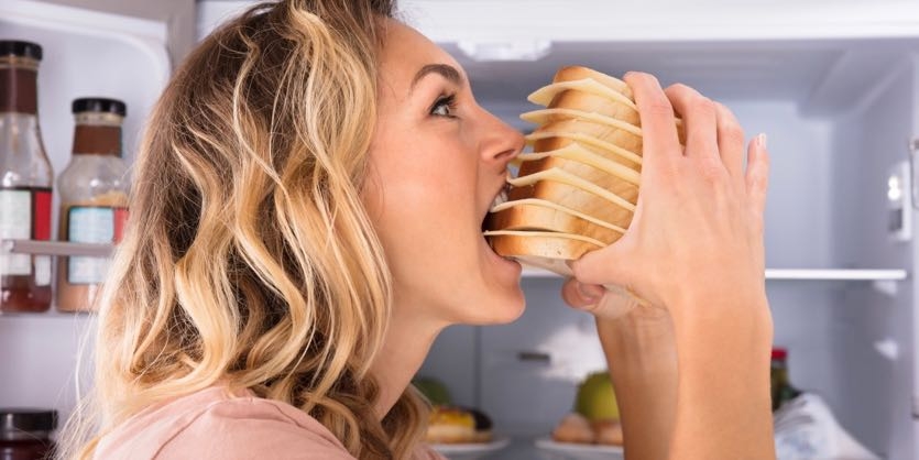 Close-up Of A Hungry Woman Eating Sandwich Near Refrigerator